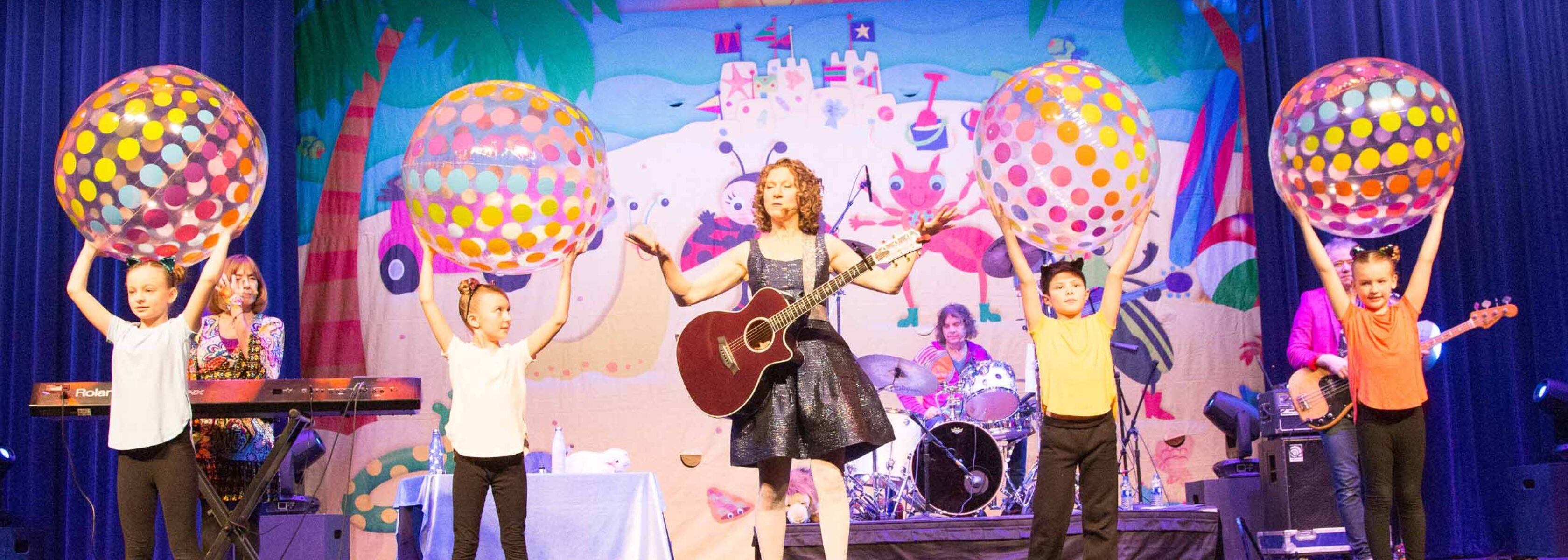 woman performing with a guitar on stage next to kids holding inflatable balls over their heads.